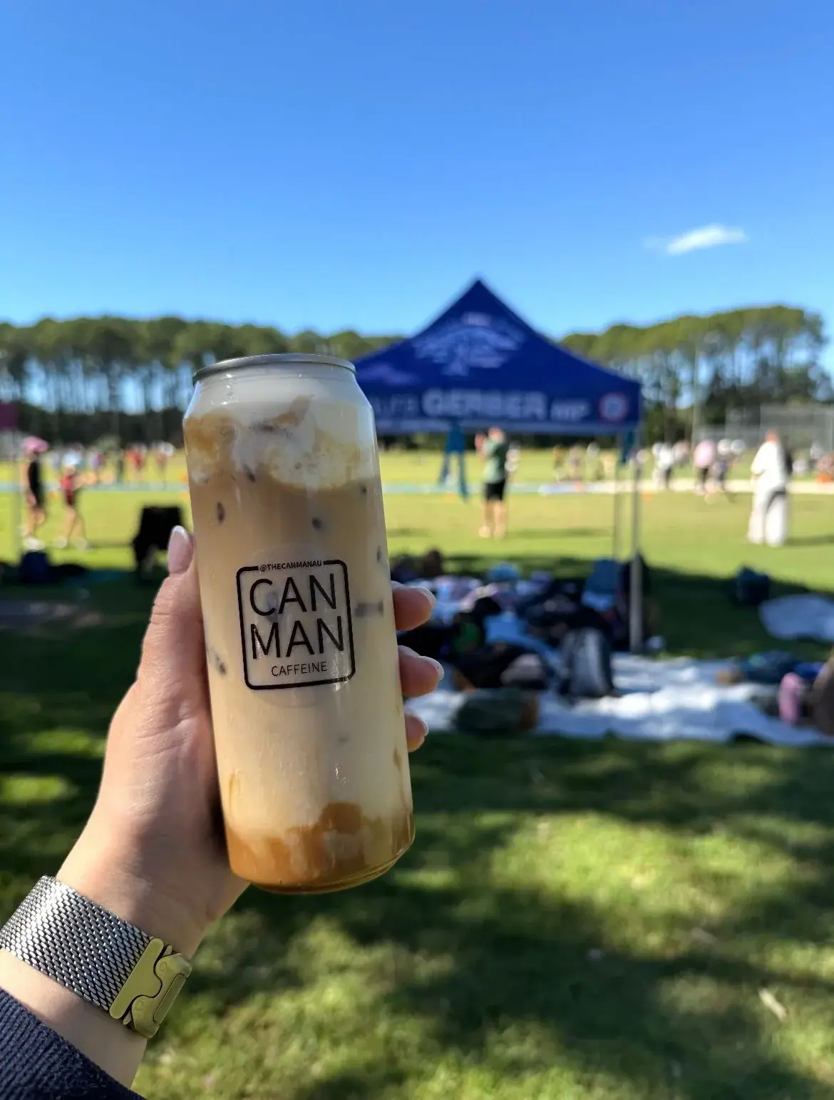 Hand holding a Can Man iced coffee sealed in a clear PET can beside a junior sports carnival sideline