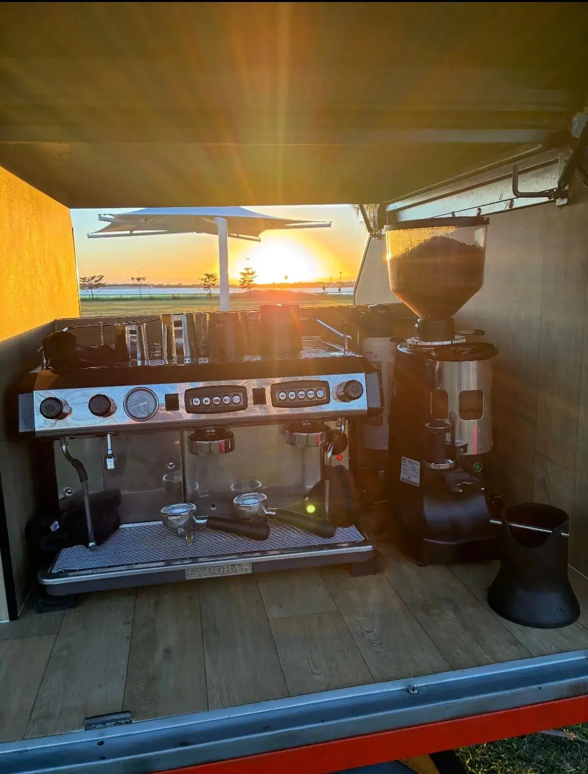 Sunrise view from inside the open gull-wing of the Can Man Coffee trailer, showing the commercial two-group espresso machine and grinder overlooking the water